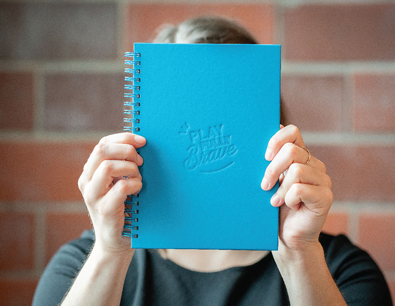 Person holding blue notebook that says "Play Fully Brave"